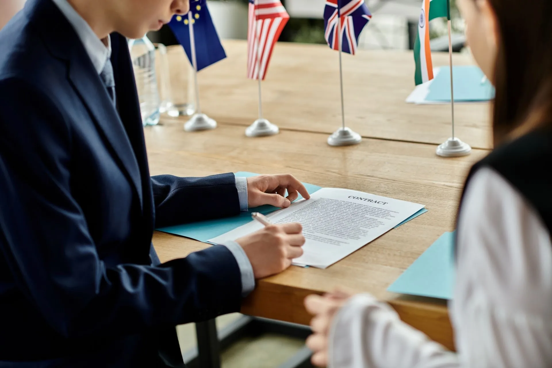 A person in a suit signing a contract at a table with international flags in the background, representing the importance of overcoming gender bias in machine translation through accurate, culturally sensitive human translation.
