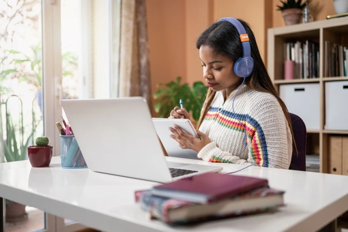 A student preparing for language testing, wearing headphones and taking notes while studying at a desk with a laptop.